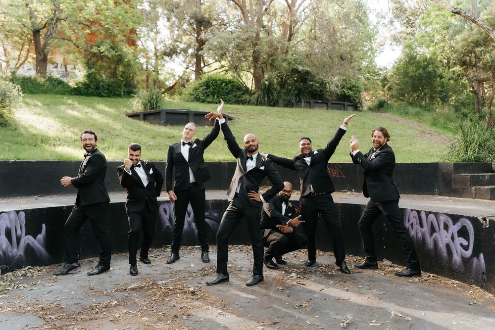 Groomsmen in black tuxedos celebrate joyfully outdoors near a graffiti wall surrounded by lush greenery.
