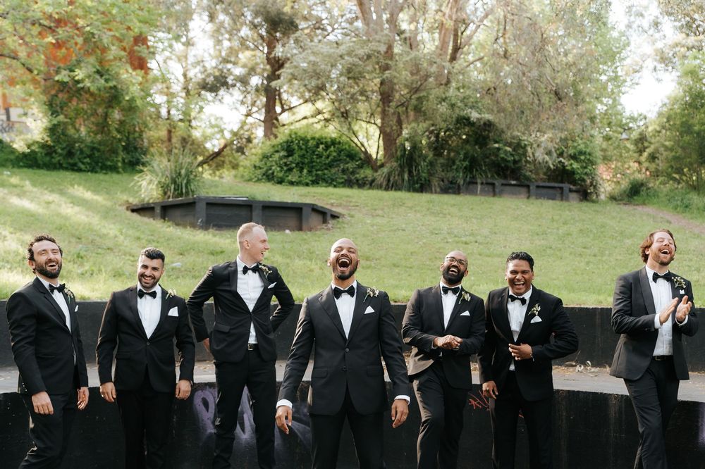 Groom and groomsmen laughing together in black tuxedos with bow ties outdoors at a wedding celebration.