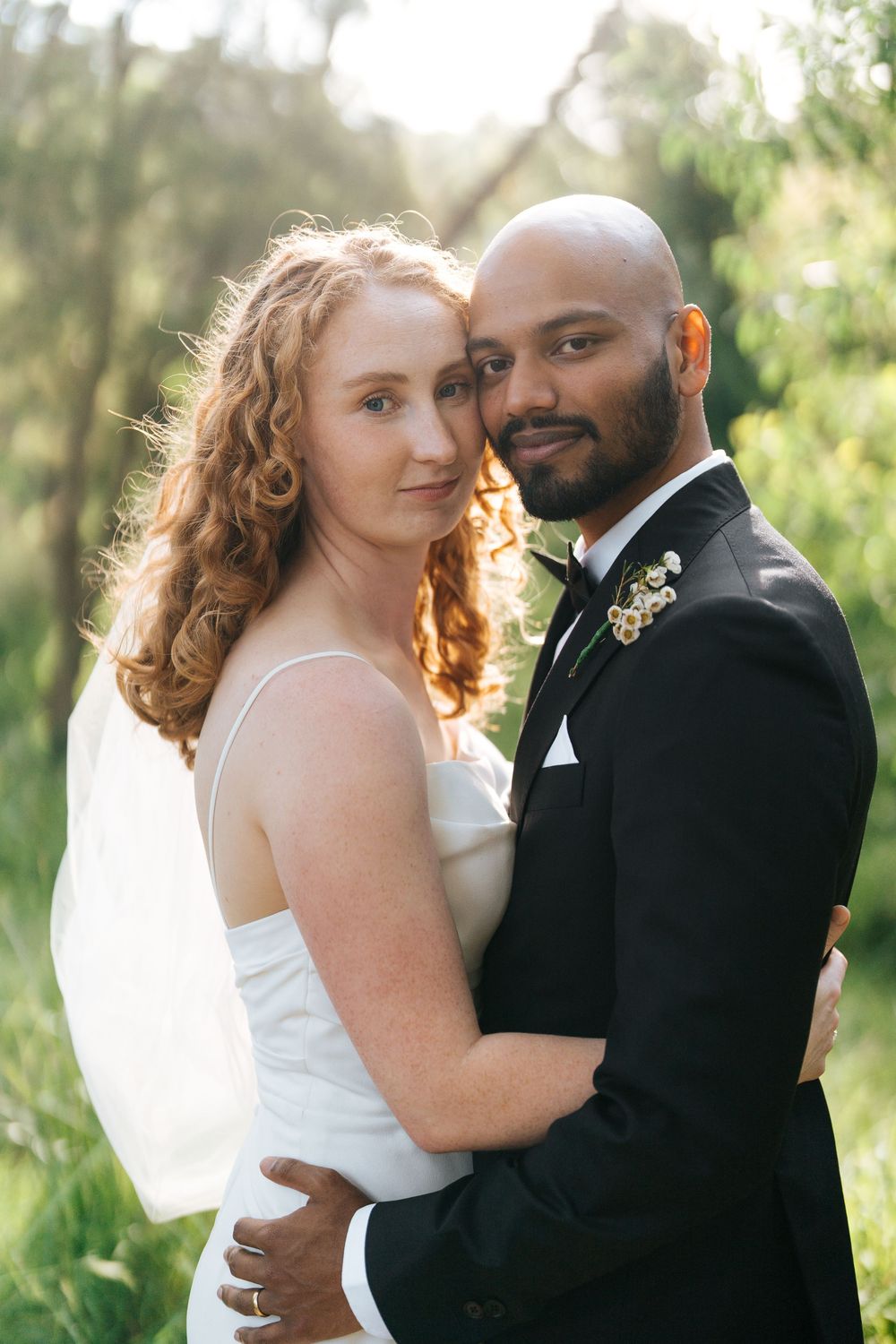 A bride and groom embrace outdoors, she in a white gown with veil, he in a black tuxedo with boutonniere.
