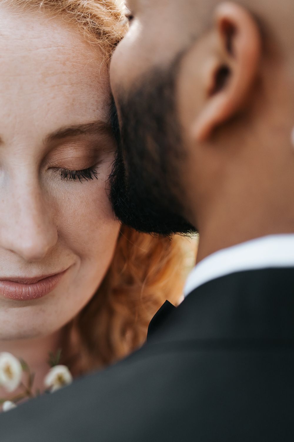Close-up of a smiling redhead bride leaning against her groom in a black suit on their wedding day.
