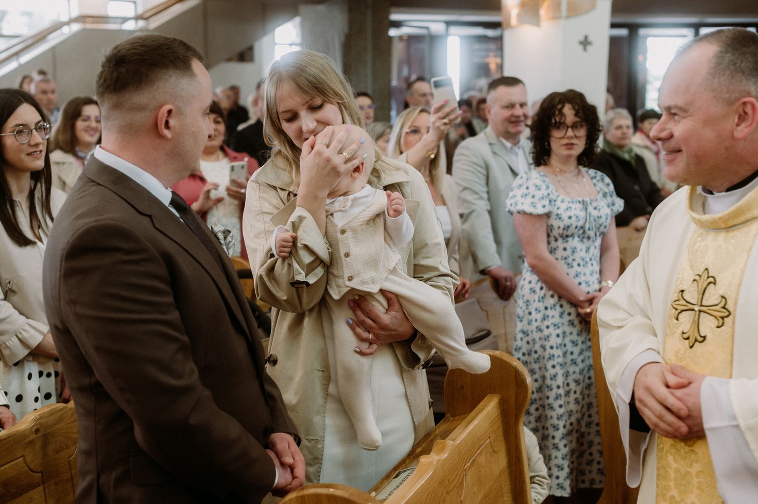 A joyful baptism ceremony with a priest, parents, and baby in a church filled with guests.