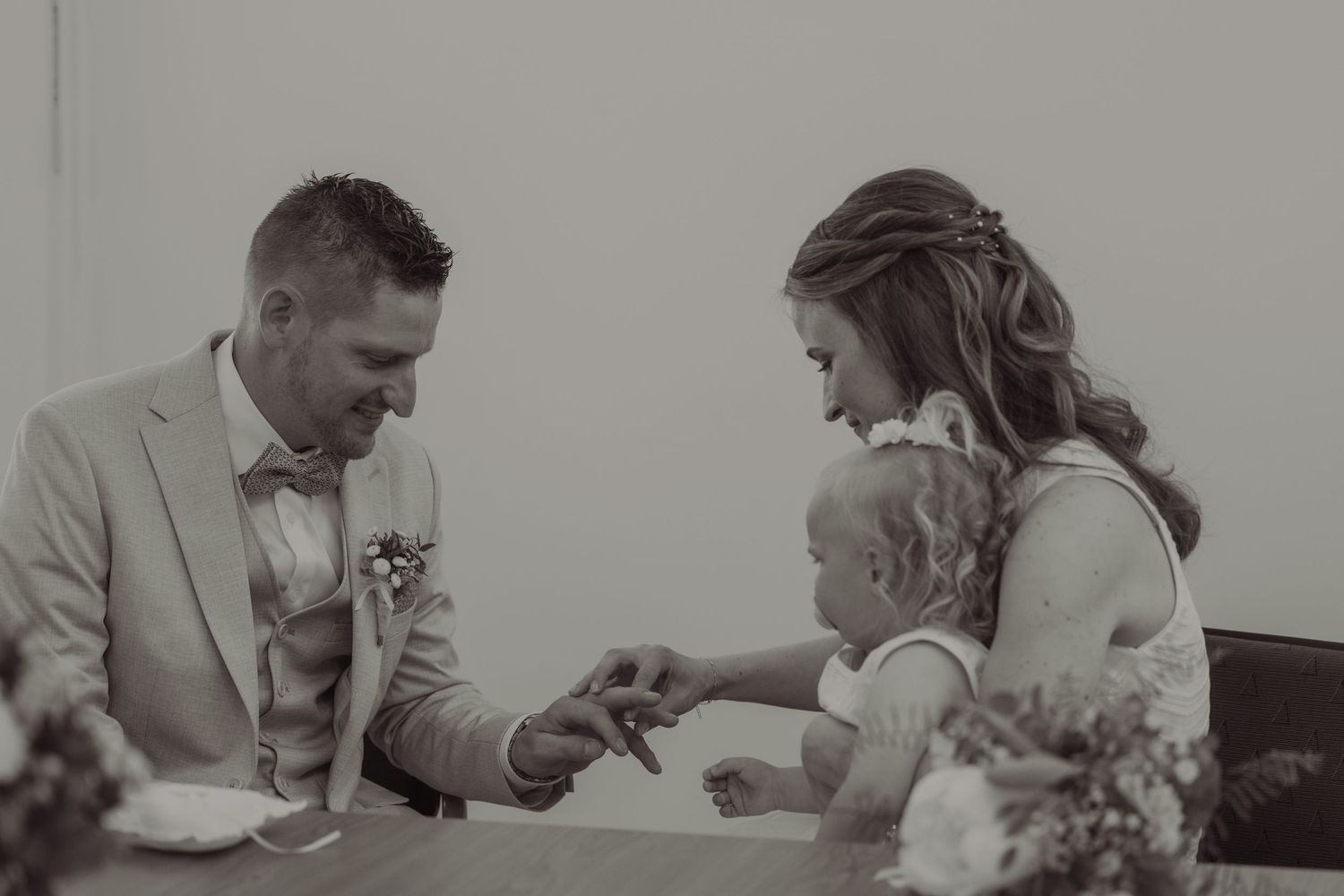 Black and white wedding photo of bride and groom signing documents with their young child at the ceremony table.