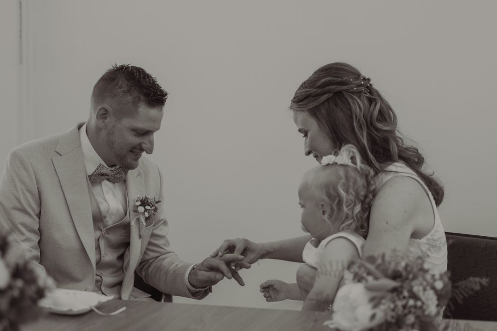 Black and white wedding photo of bride and groom signing documents with their young child at the ceremony table.