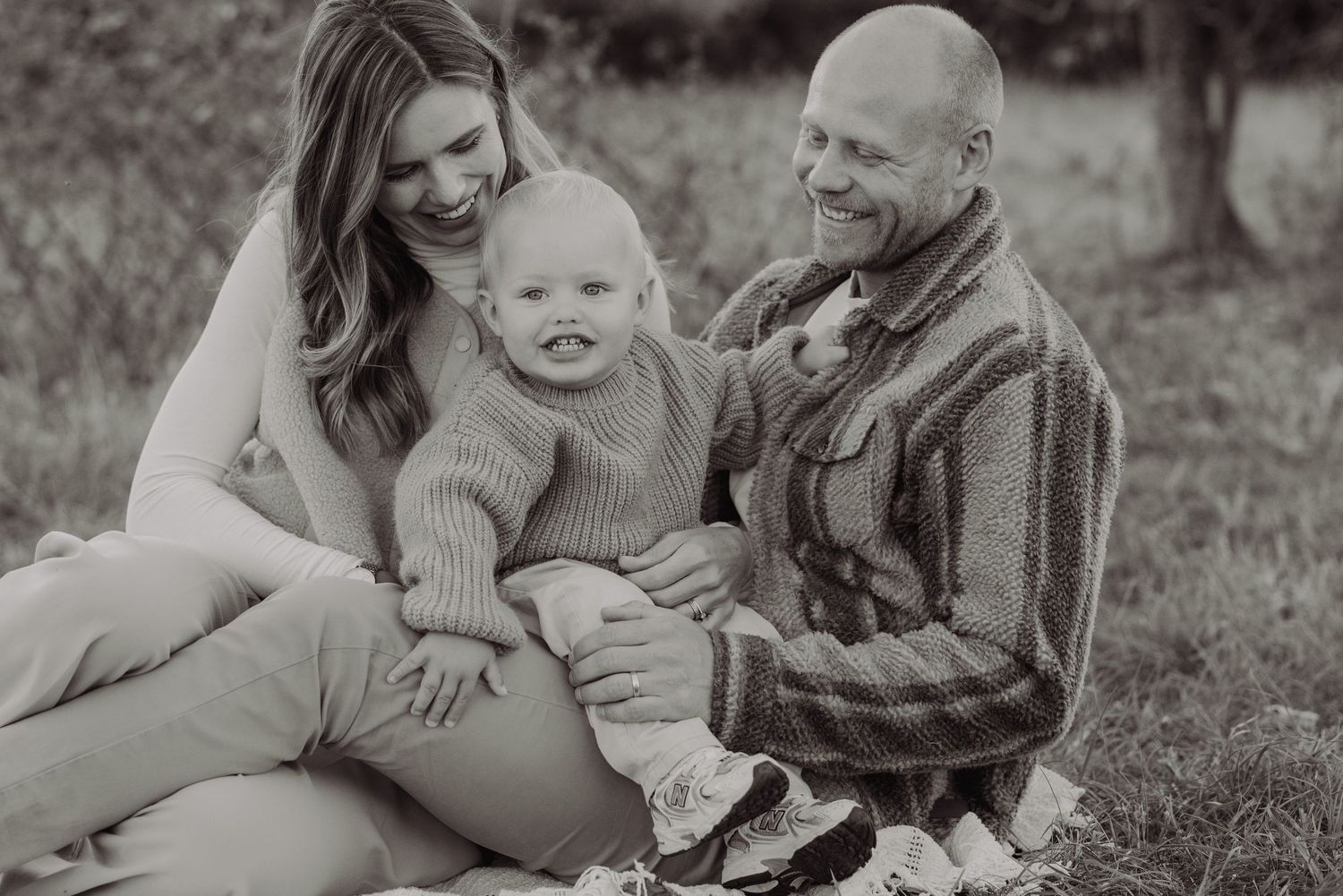 Black and white family photo of smiling parents sitting outdoors with their happy baby in a knit sweater.
