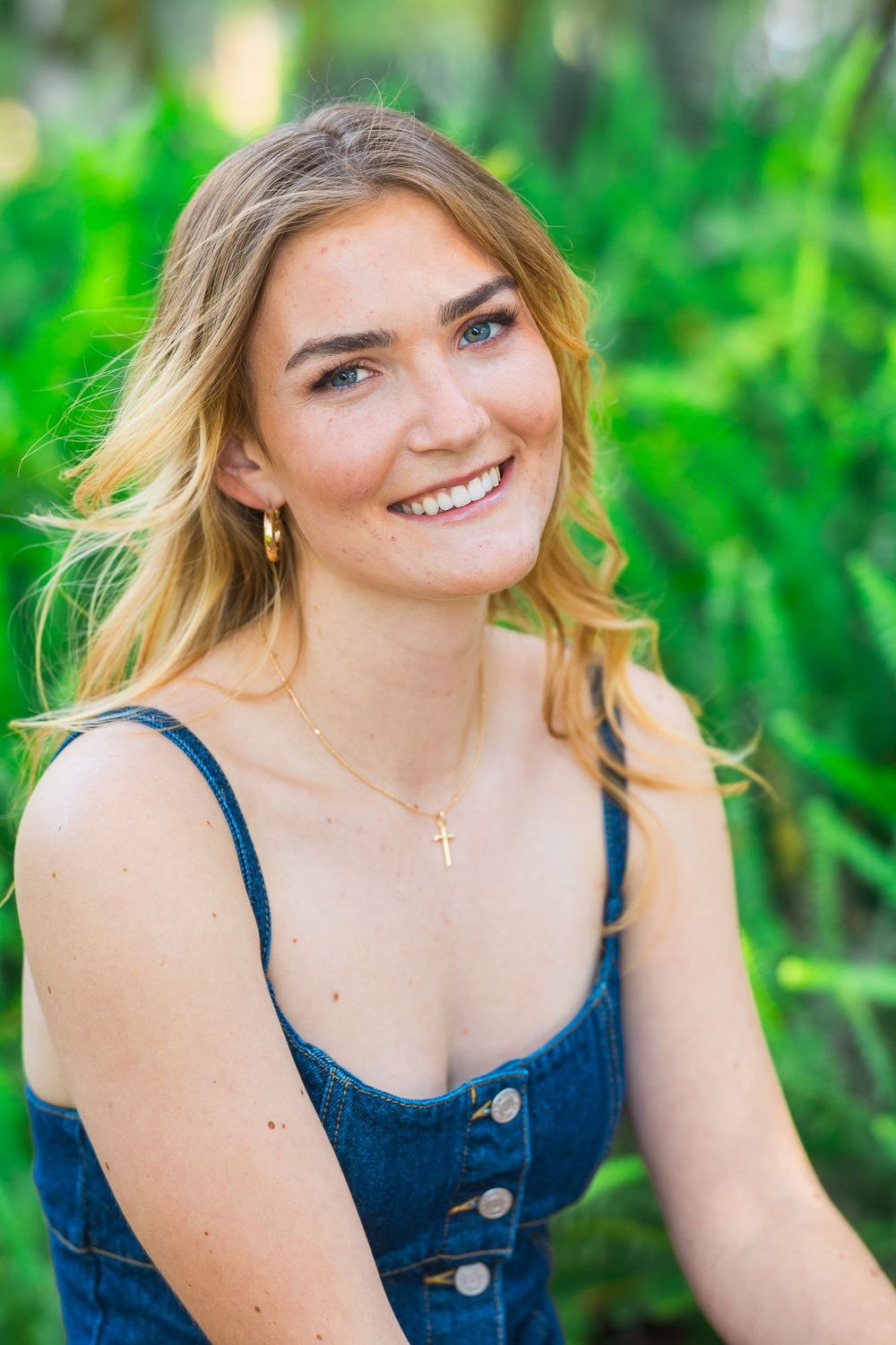 A portrait of someone in a blue denim dress smiling outdoors with lush green foliage in the background.