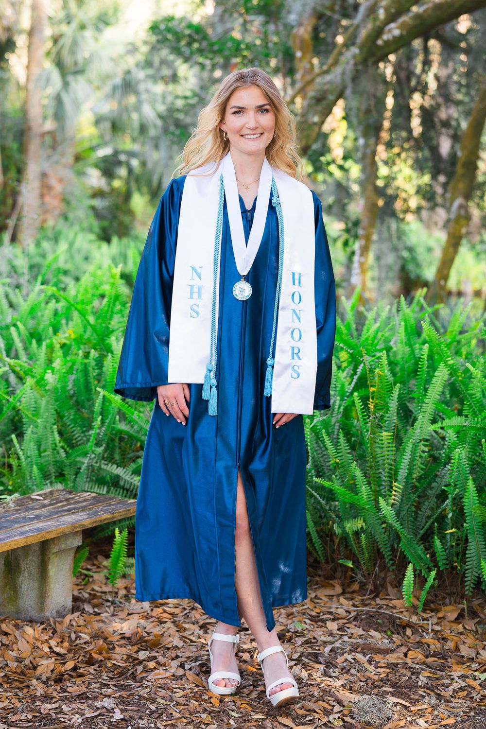 Graduate in blue and white academic regalia standing among tropical ferns.