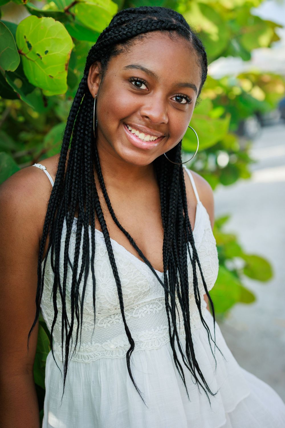 Portrait of someone with long braids wearing a white dress and hoop earrings against green foliage.