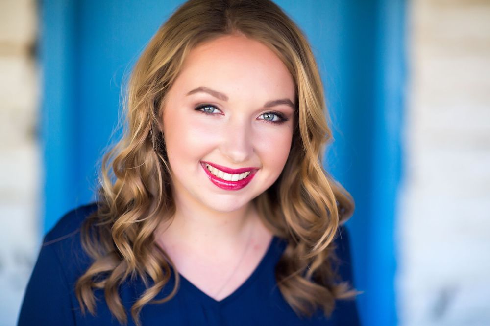 Professional portrait of someone in a navy v-neck top with wavy blonde hair and bright red lipstick against a blue background.