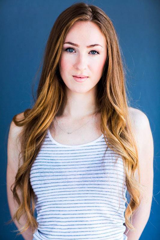 Young woman with long wavy brown hair wearing a striped tank top poses against blue background.