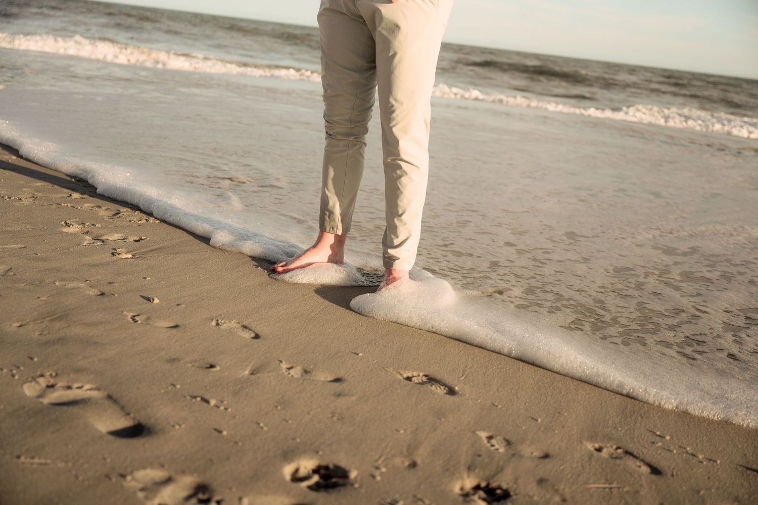 Bare feet standing in ocean waves on a sandy beach at sunset.