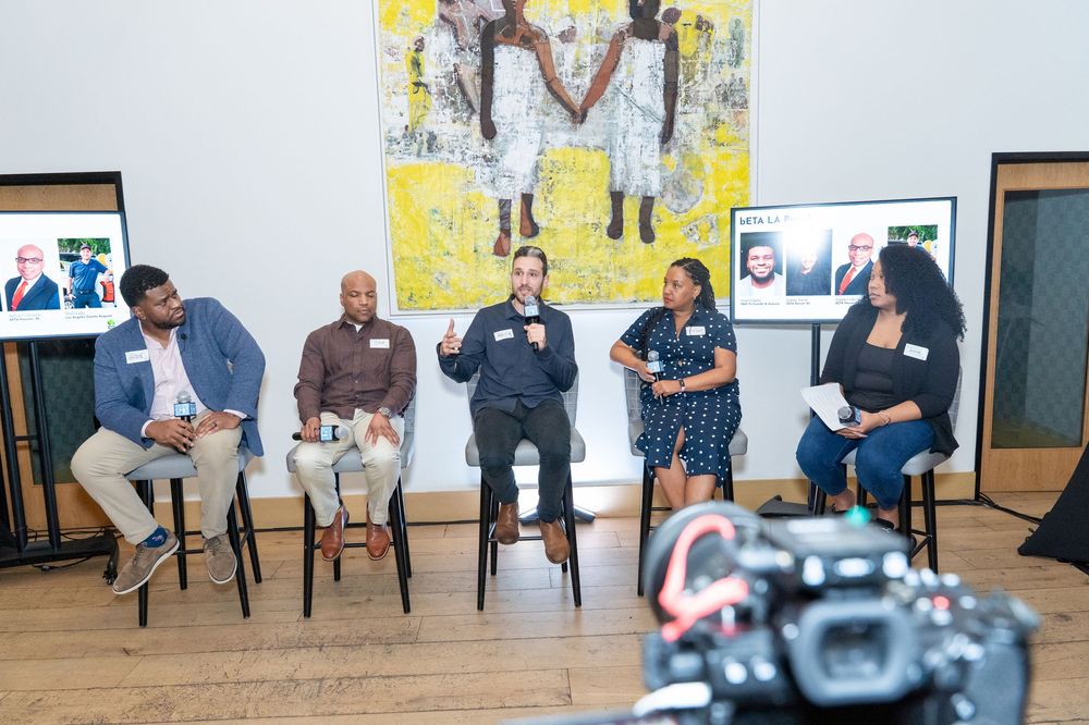Panel discussion with five diverse speakers seated before a colorful painting at an indoor event.