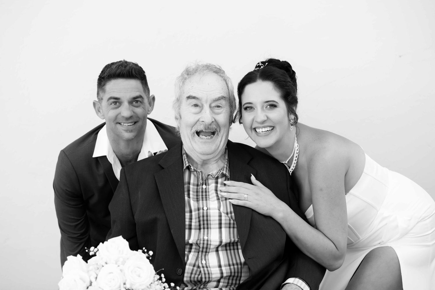Black and white wedding photo of bride and groom posing with elderly man, all smiling joyfully.