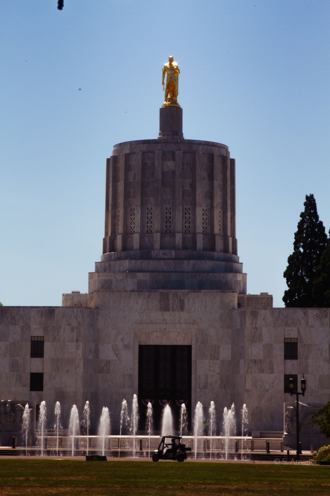 Oregon State Capitol building with golden pioneer statue atop rotunda, fountains in foreground under clear blue sky.