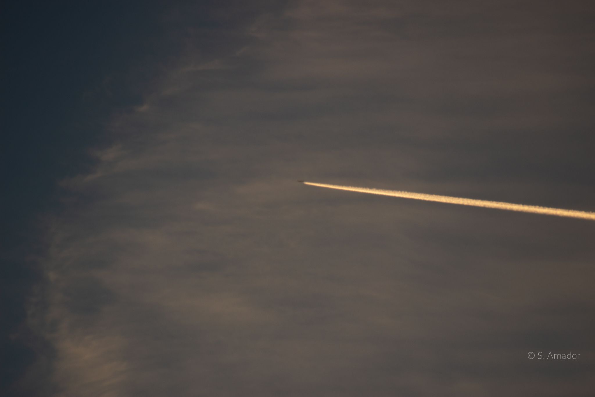 A golden contrail streaks across a dramatic cloudy sky at sunset, left by a distant aircraft flying overhead.