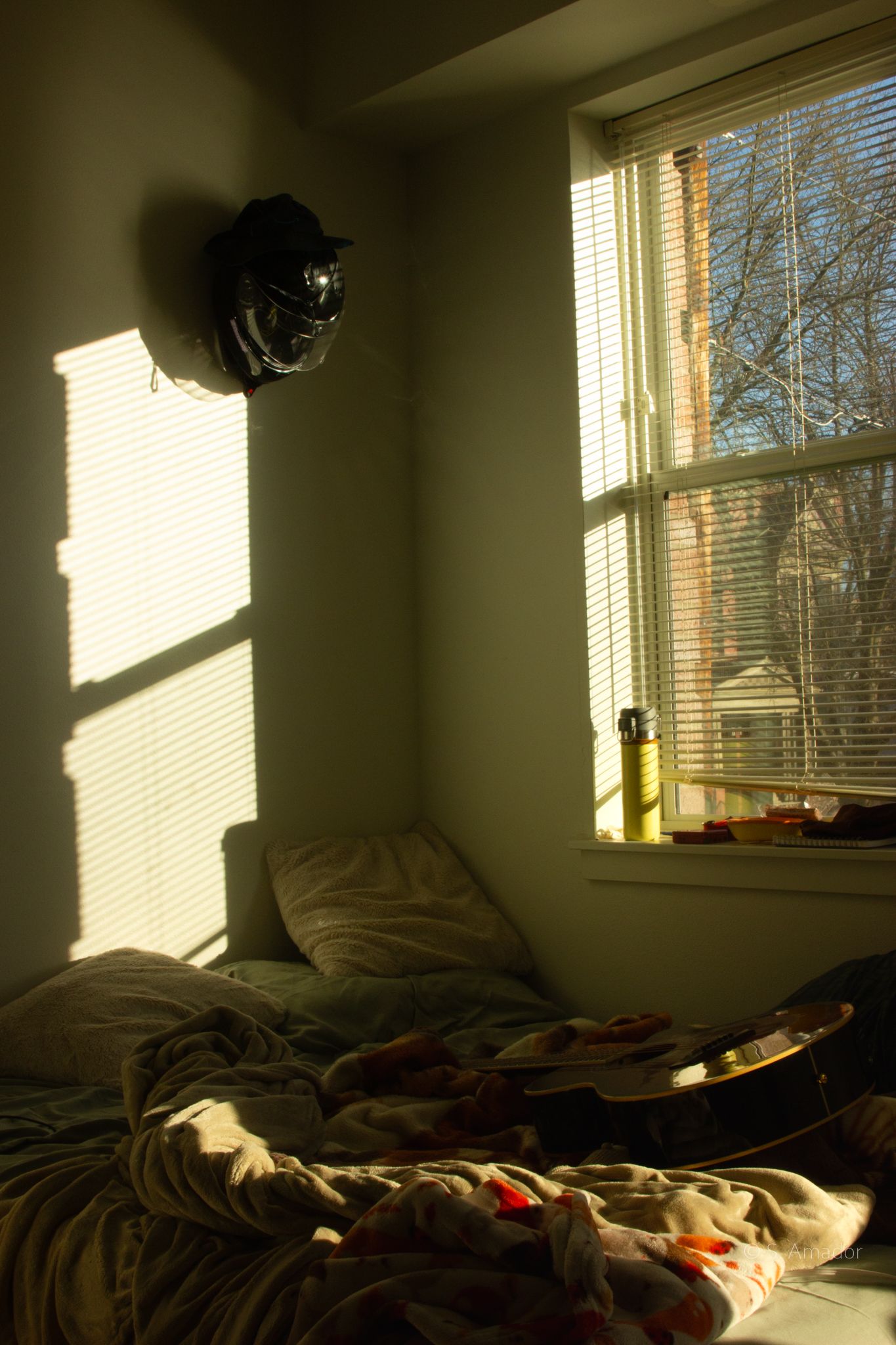 Sunlight streams through window blinds onto unmade bed with helmet mounted on wall in cozy bedroom.