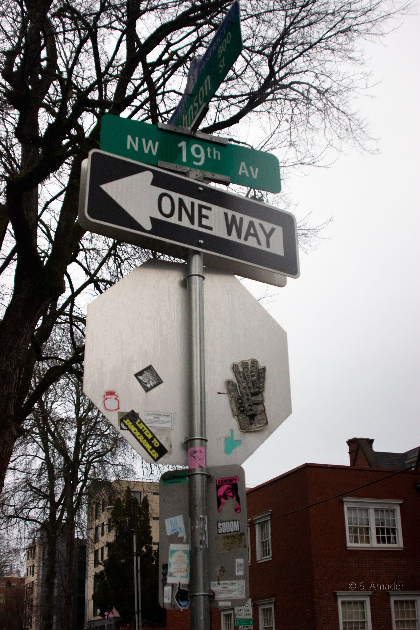 NW 19th Avenue one way street sign on pole with stickers, bare trees and brick building background.