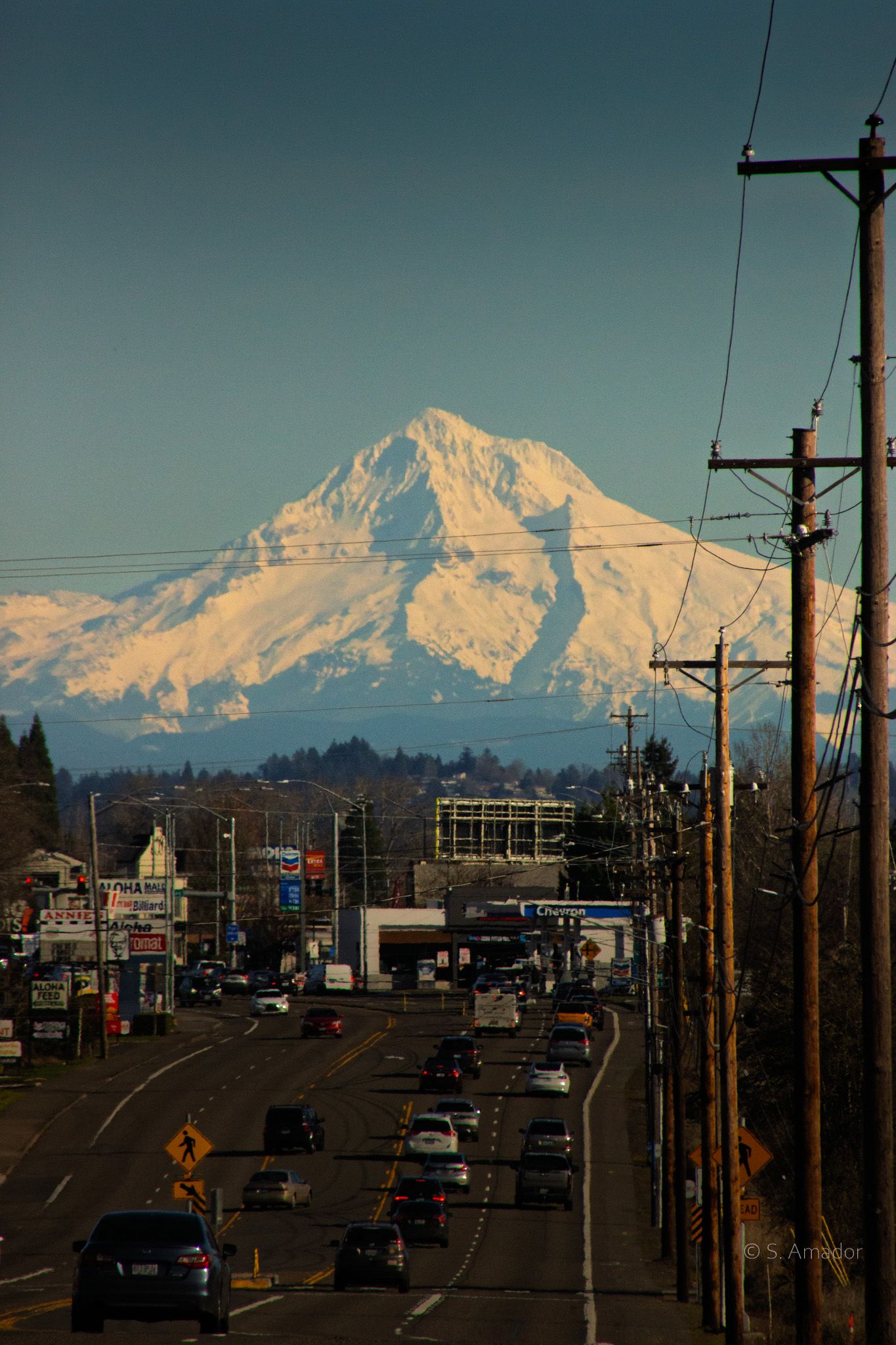 Snow-capped Mount Hood towers over a busy urban street with traffic and power lines in Portland.