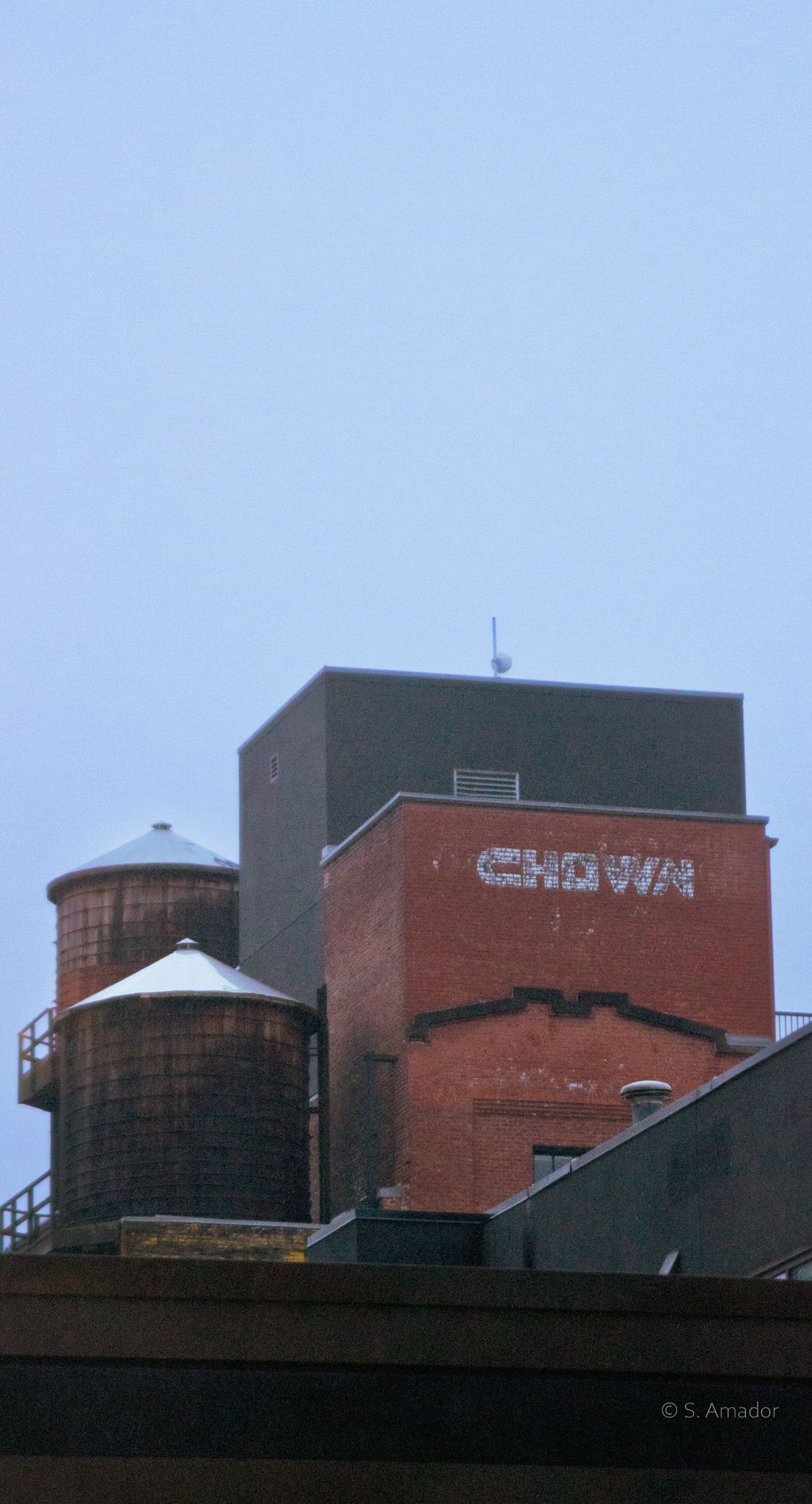 Historic red brick building rooftop with a wooden water tower and faded CHOWN sign against gray sky.