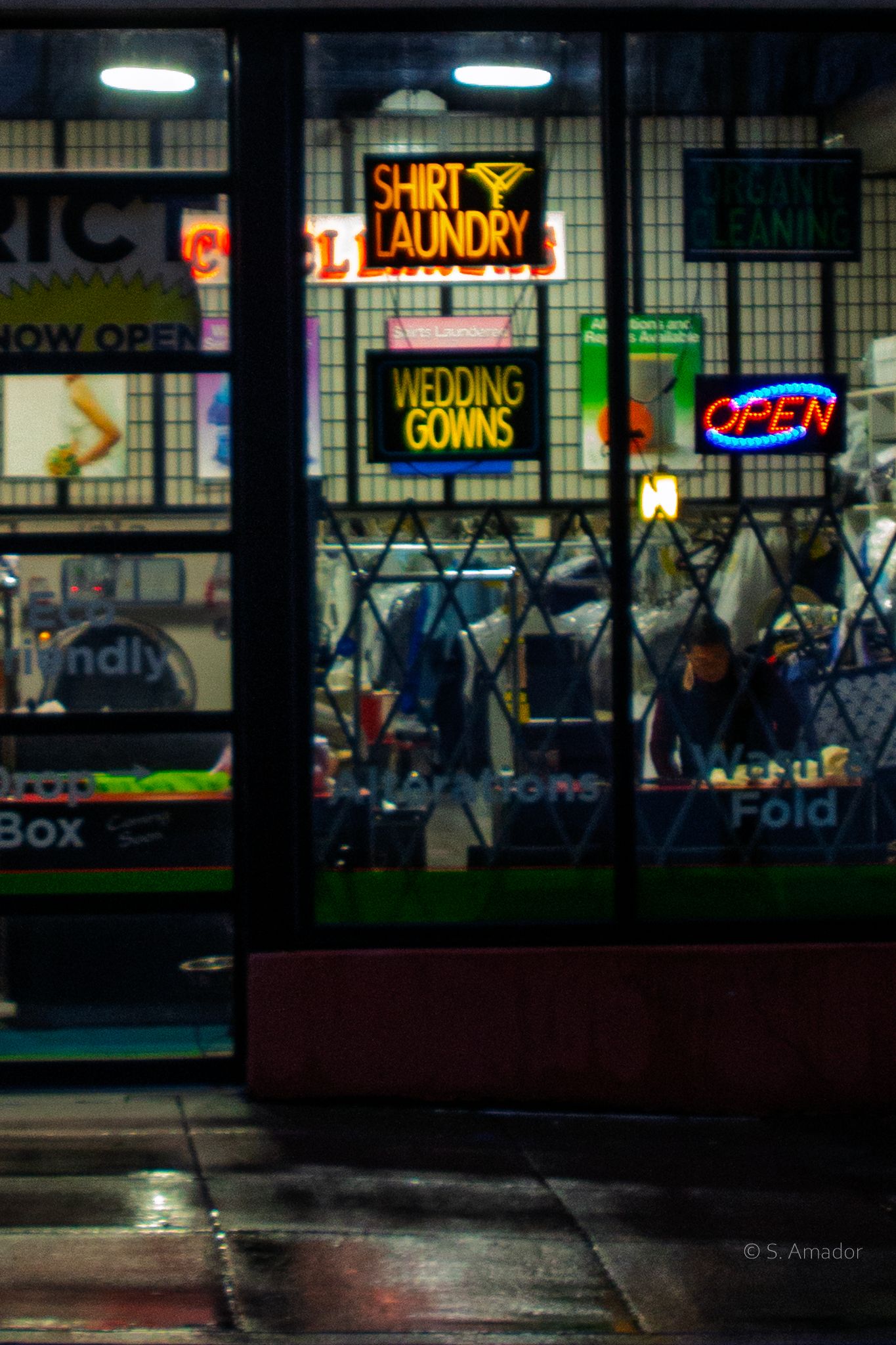 Neon signs for shirt laundry and wedding gowns glow in a storefront window at night on a wet street.