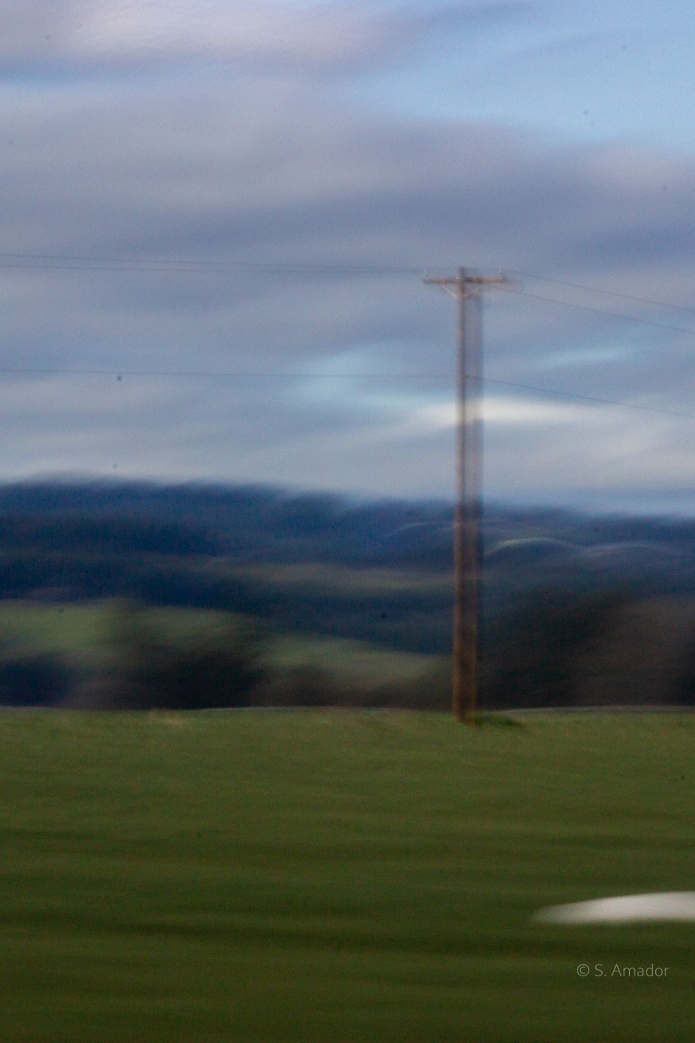 A blurred motion shot of a rustic utility pole standing in a green field with rolling hills in background.
