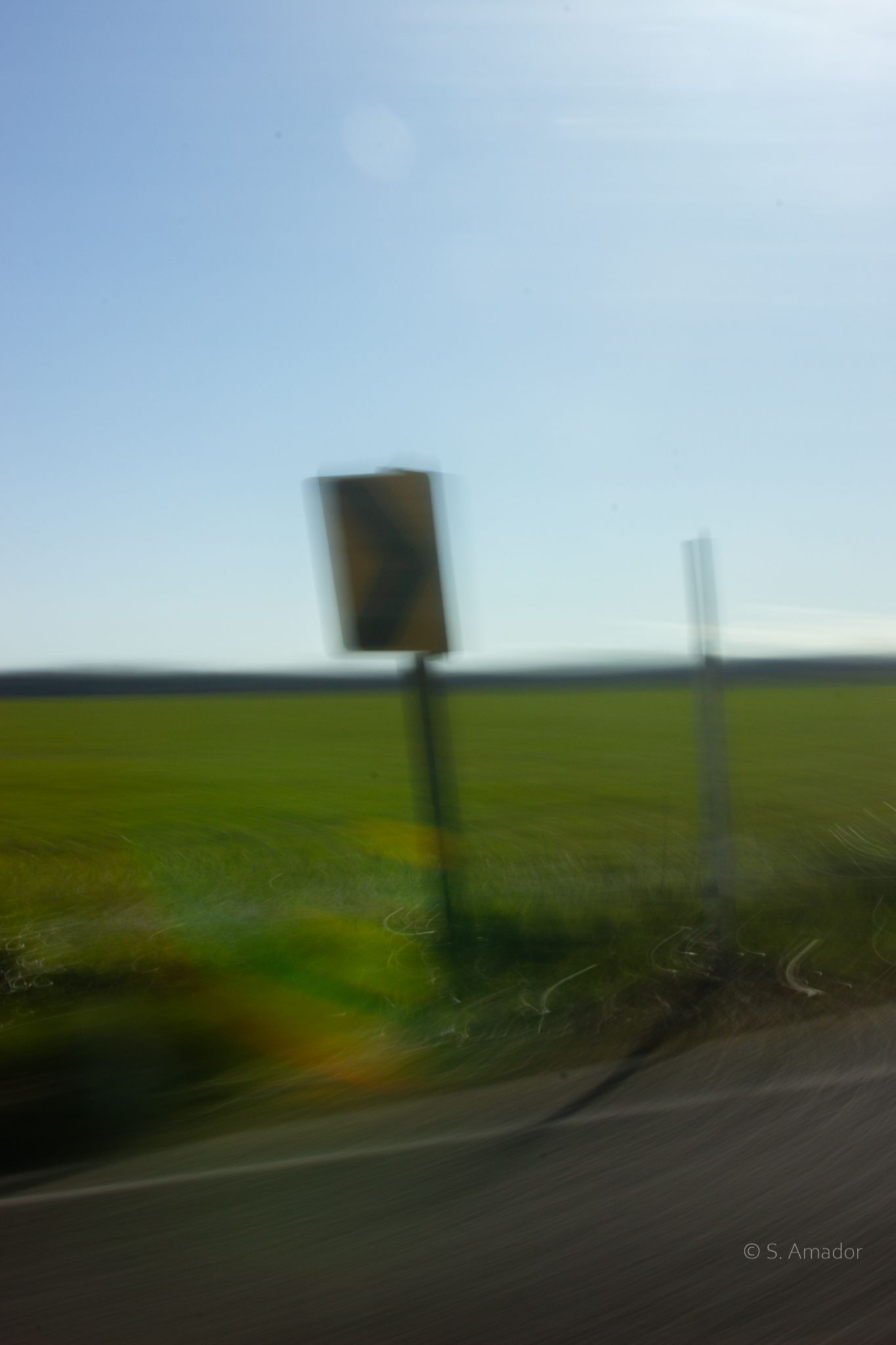 Blurred motion shot of a roadside warning sign beside a green field under a clear blue sky.