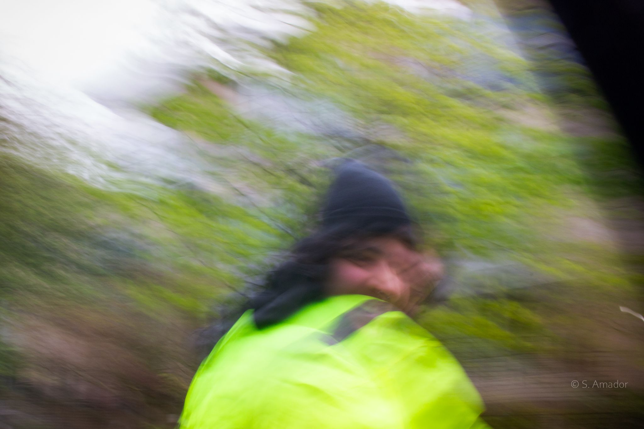 Blurred motion photo of person in yellow high-visibility jacket and dark hat moving through green foliage.