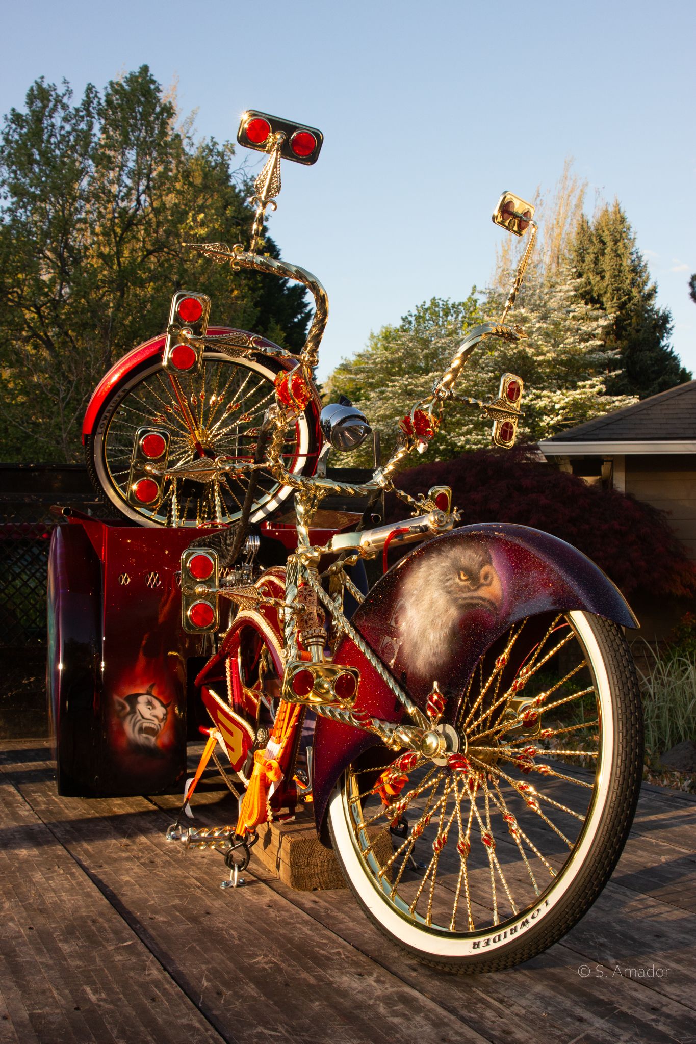 Elaborately customized red and gold lowrider bicycle with eagle design displayed on wooden deck outdoors.