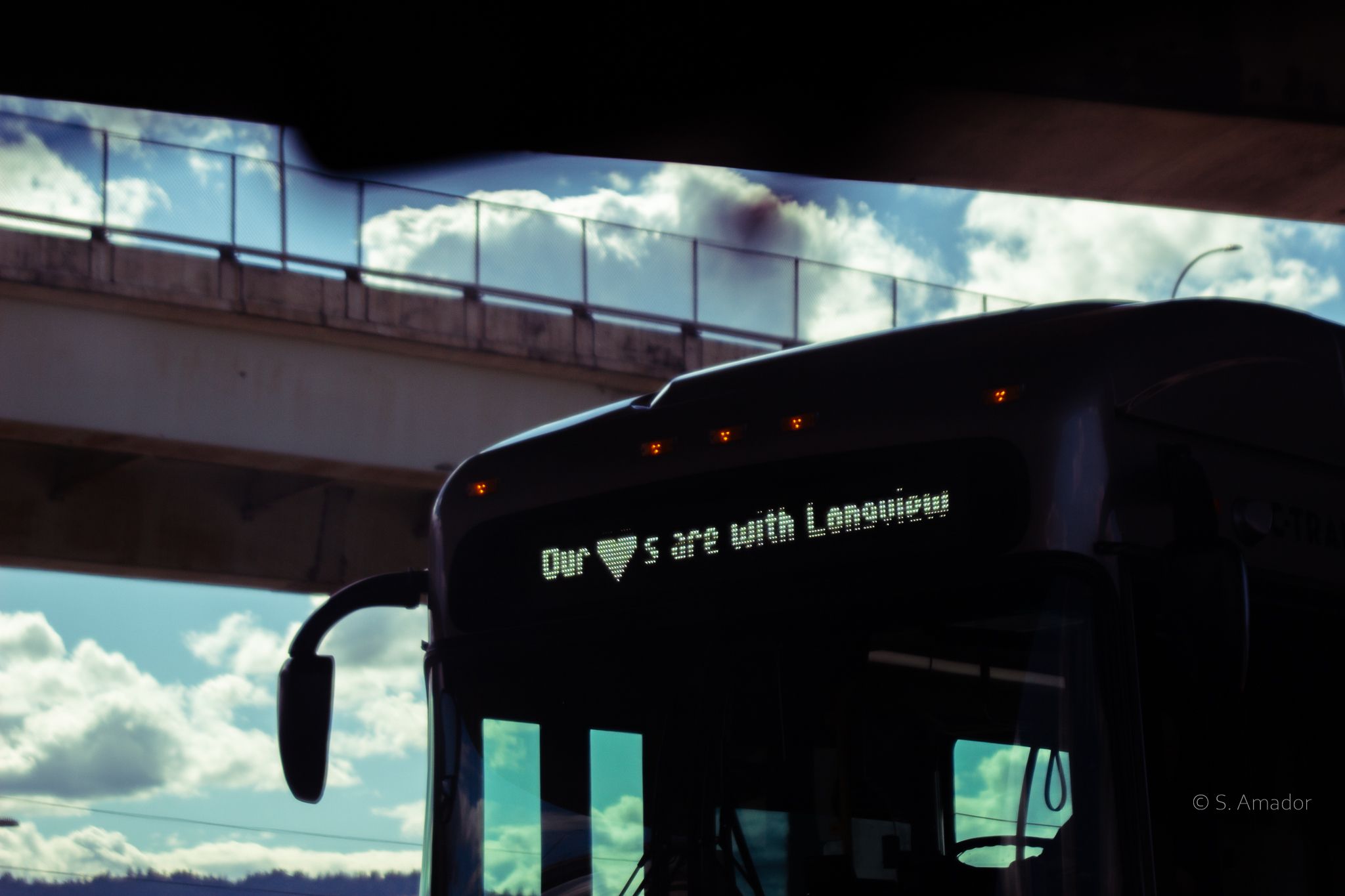 City bus displaying Our Hearts are with Longview message beneath an overpass against dramatic cloudy sky.