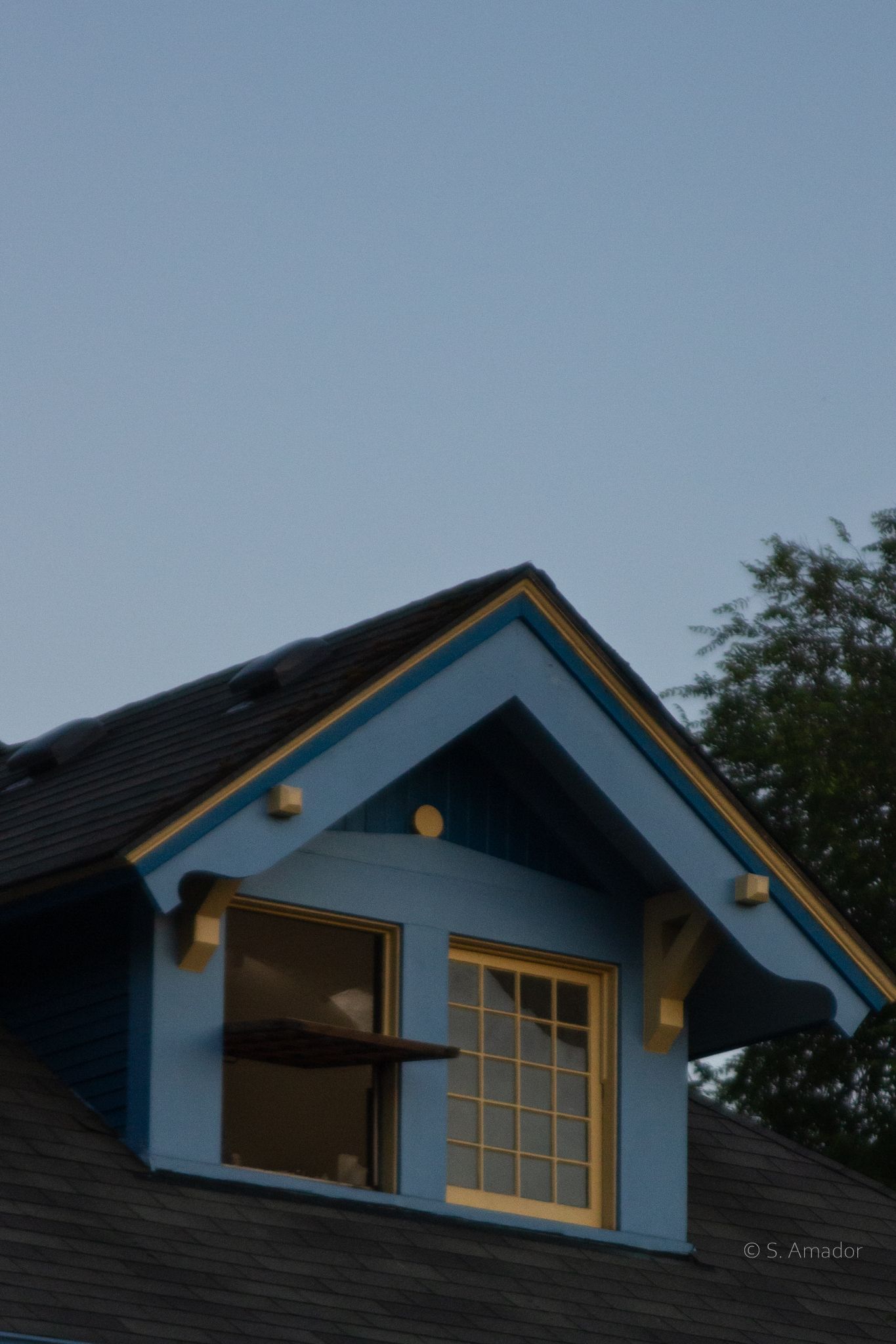 Blue dormer window on residential rooftop at dusk with yellow trim and warm interior glow.