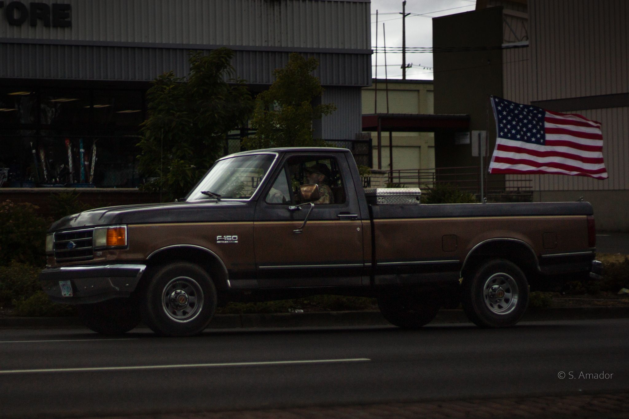 Brown Ford F-150 pickup truck with American flag driving on a street in a suburban area.