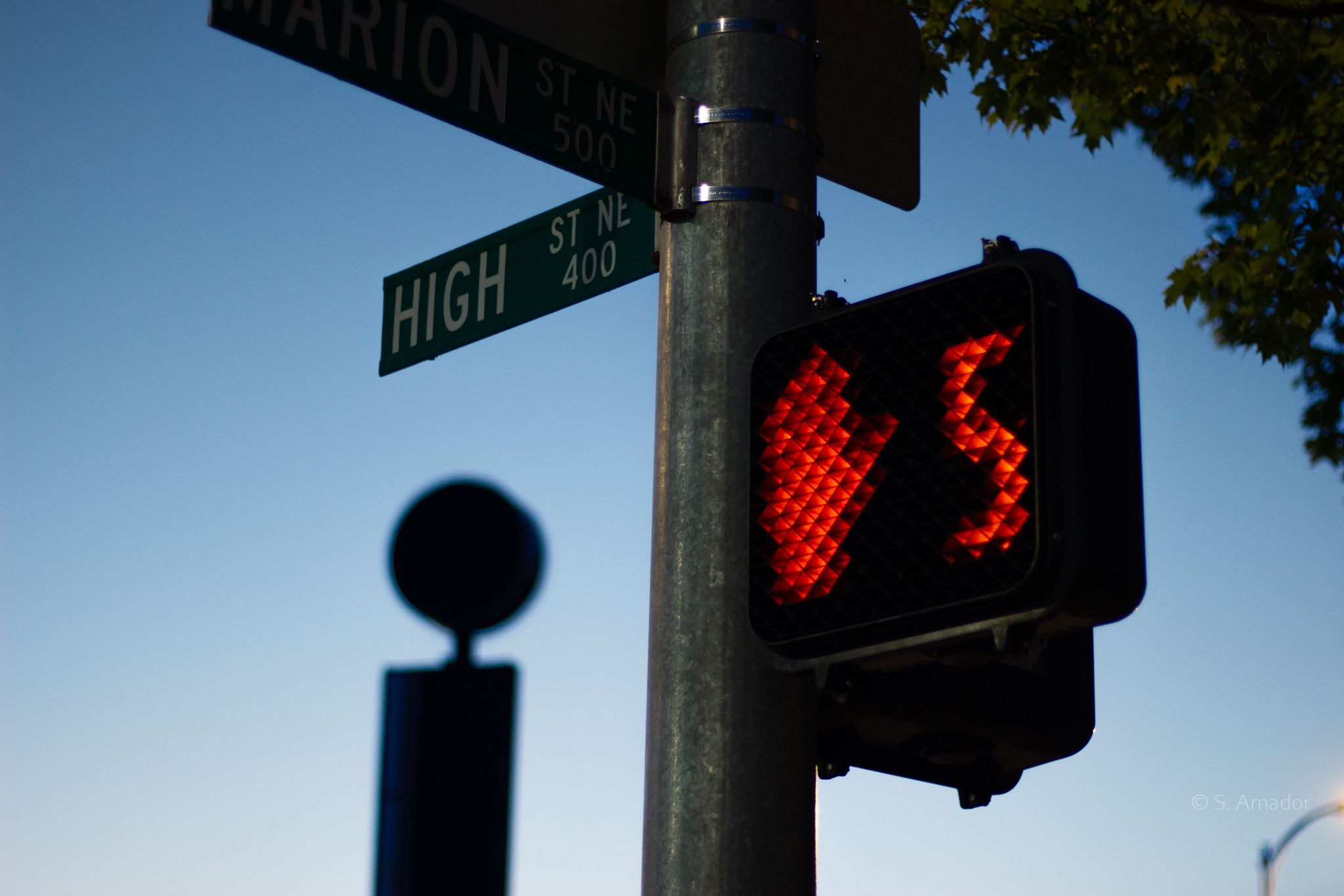 Red pedestrian stop signal with countdown timer at High St NE and Marion St intersection against clear blue sky.