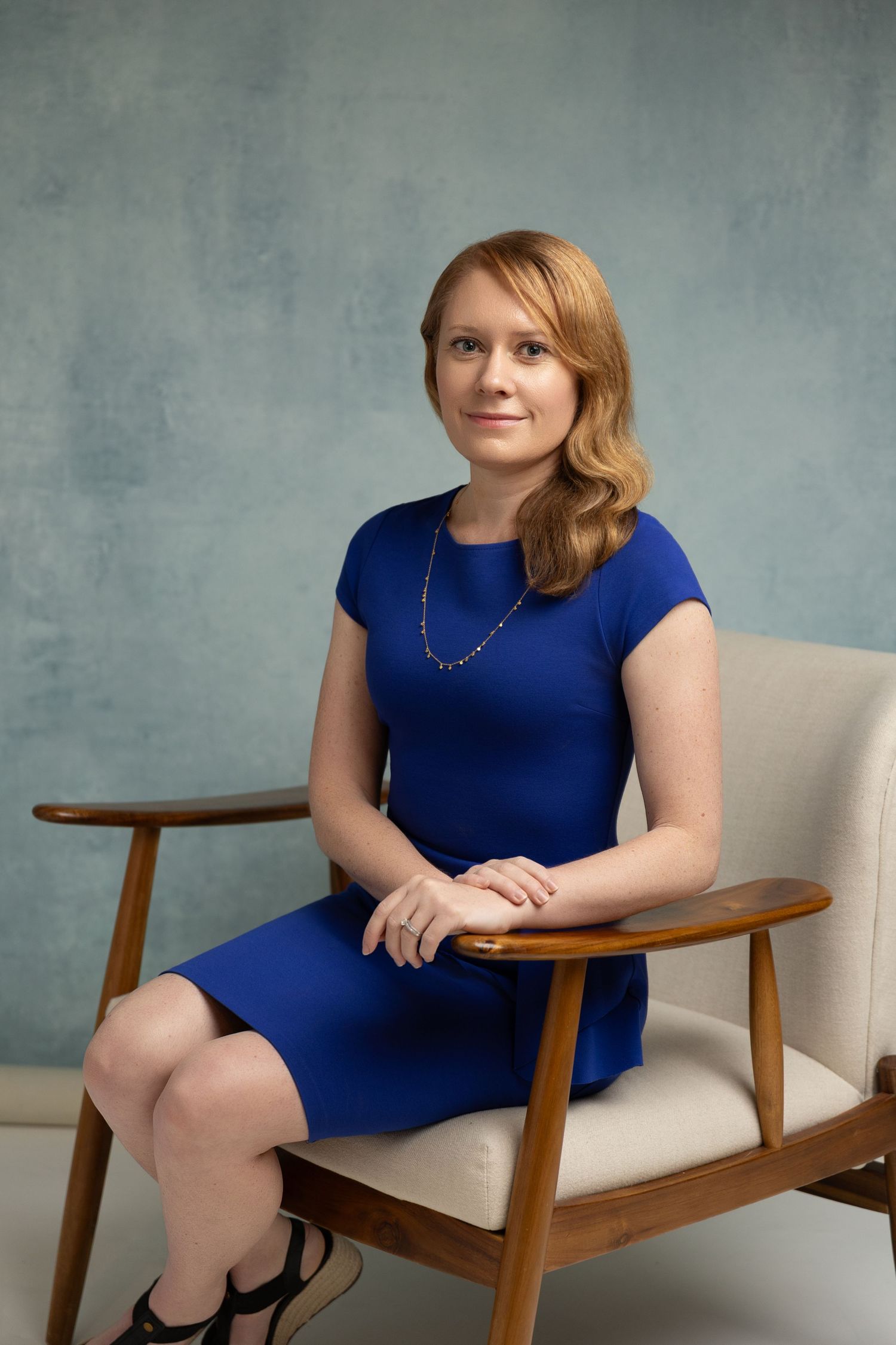 Professional woman in blue dress seated elegantly in a mid-century modern chair against a blue-gray backdrop.
