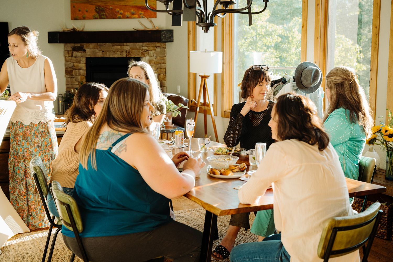 Women gathered around a dining table sharing a meal in a cozy stone fireplace room with natural light.