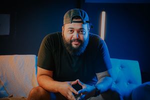 A bearded man wearing a black t-shirt and backward cap sits on a couch, smiling in blue-tinted ambient lighting.