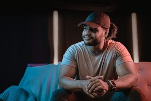 Young man in red cap and gray t-shirt sits relaxed, smiling in moody blue and pink neon lit room.
