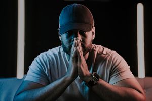 Man in white t-shirt and cap praying with hands together in dramatic blue neon lighting.