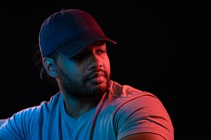 Bearded man in blue cap and t-shirt under dramatic red and blue neon lighting against dark background.