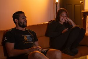 Two people sitting on a couch in warm ambient lighting, smiling and relaxed in casual black outfits.