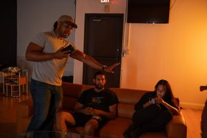 Man standing and pointing at two people sitting on a brown couch in a dimly lit room.