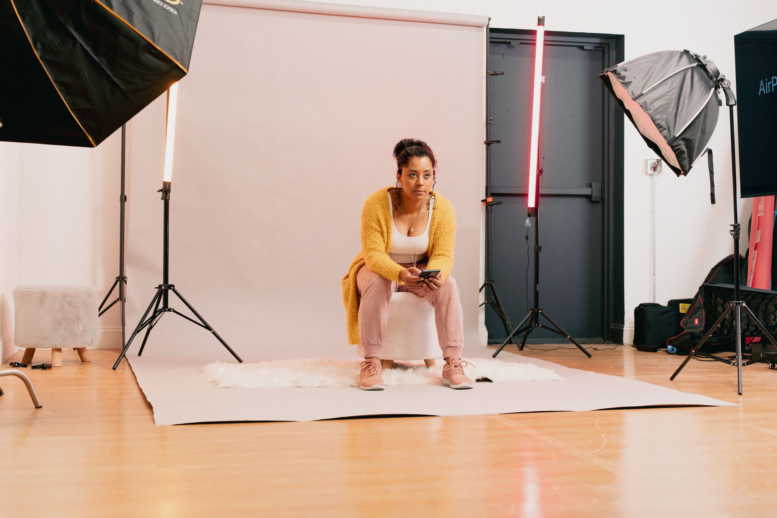 Woman in yellow cardigan sitting on stool in professional photo studio with lighting equipment and white backdrop.