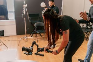 A female videographer operates a professional camera gimbal stabilizer on a film set with studio lighting equipment.