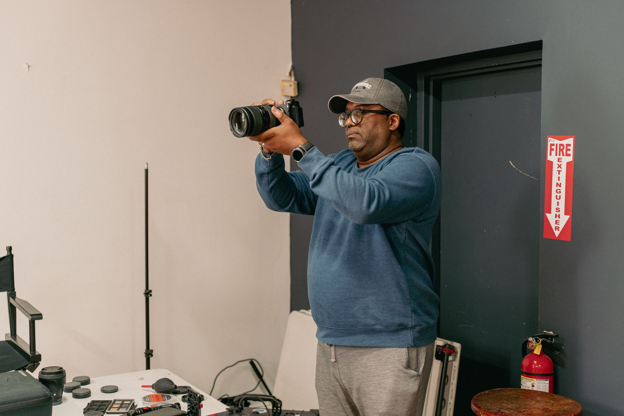 A man wearing a gray cap and blue hoodie photographs with a DSLR camera in a photography studio.