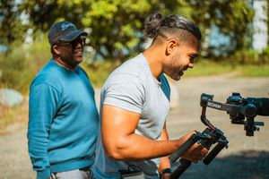 Two men outdoors, one holding a DJI Ronin camera gimbal stabilizer, filming on a sunny day near green trees.