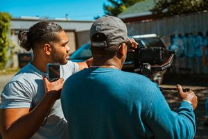 Two men collaborating on a video shoot outdoors, one holding a smartphone and the other operating a professional camera.