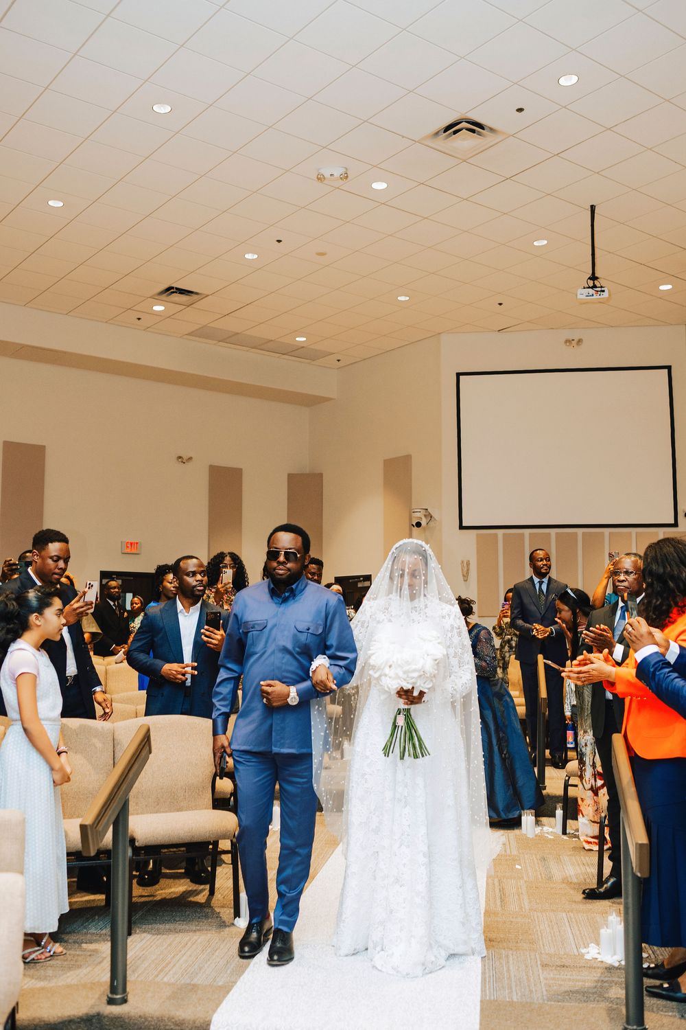 Groom in blue suit escorts veiled bride in white lace gown down church aisle during wedding ceremony.