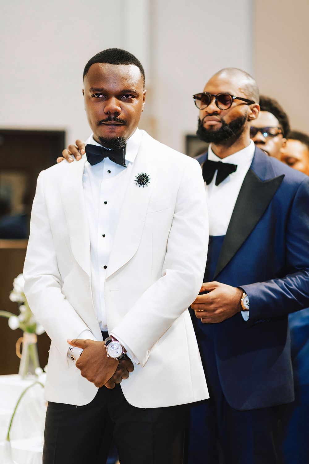 Groom in white tuxedo jacket with bow tie stands at altar, groomsmen in navy suits behind him.