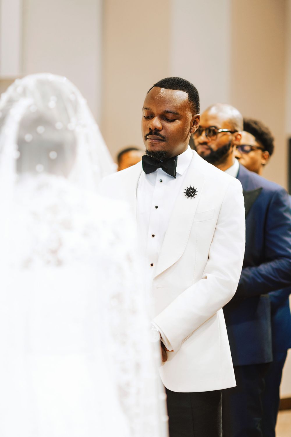 Groom in white tuxedo with black bow tie gazes at bride in elegant veil during wedding ceremony.