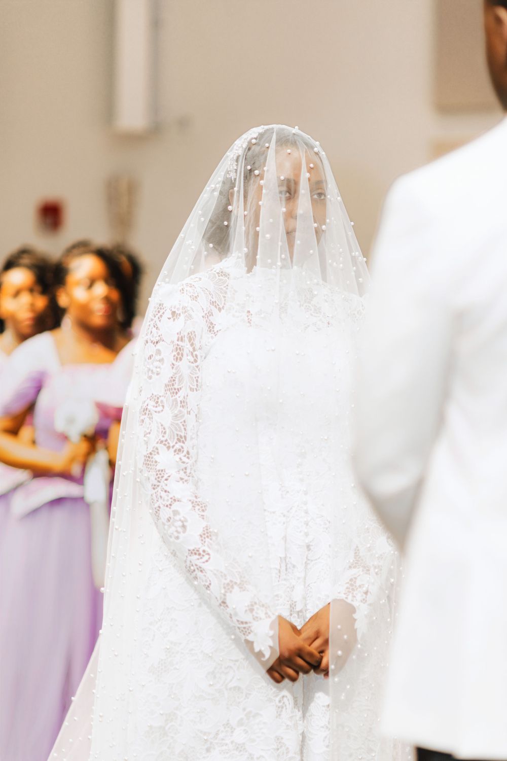 Bride in lace wedding gown and pearl-adorned veil stands at church altar with bridesmaids in lavender dresses.