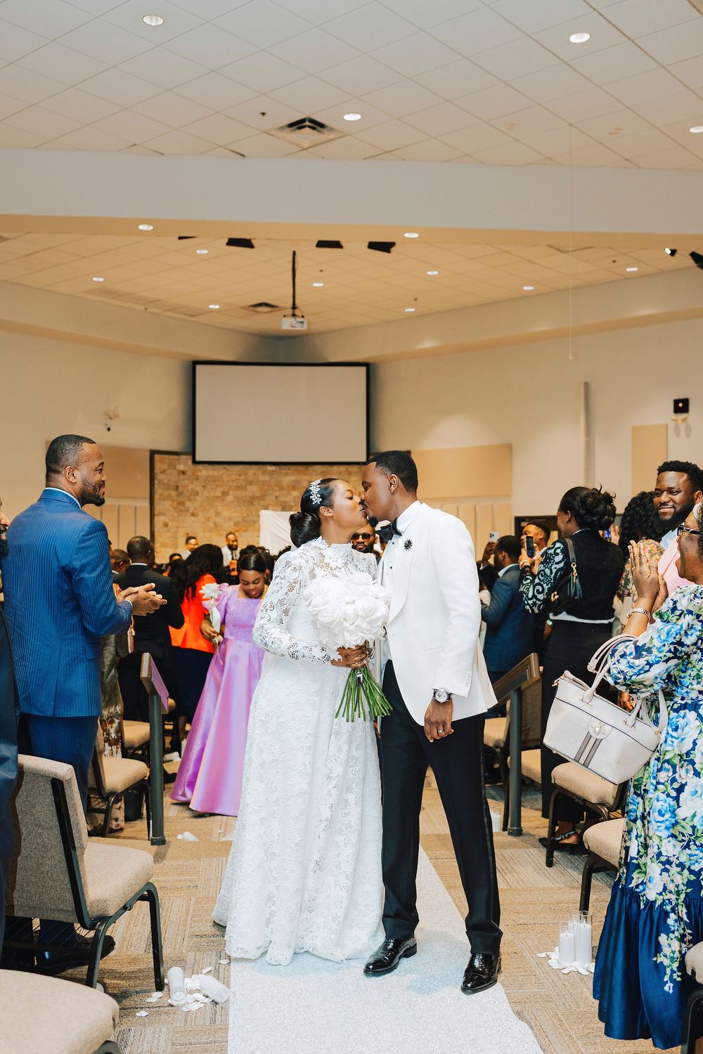 Bride and groom share a first kiss at wedding ceremony while guests applaud in decorated church venue.