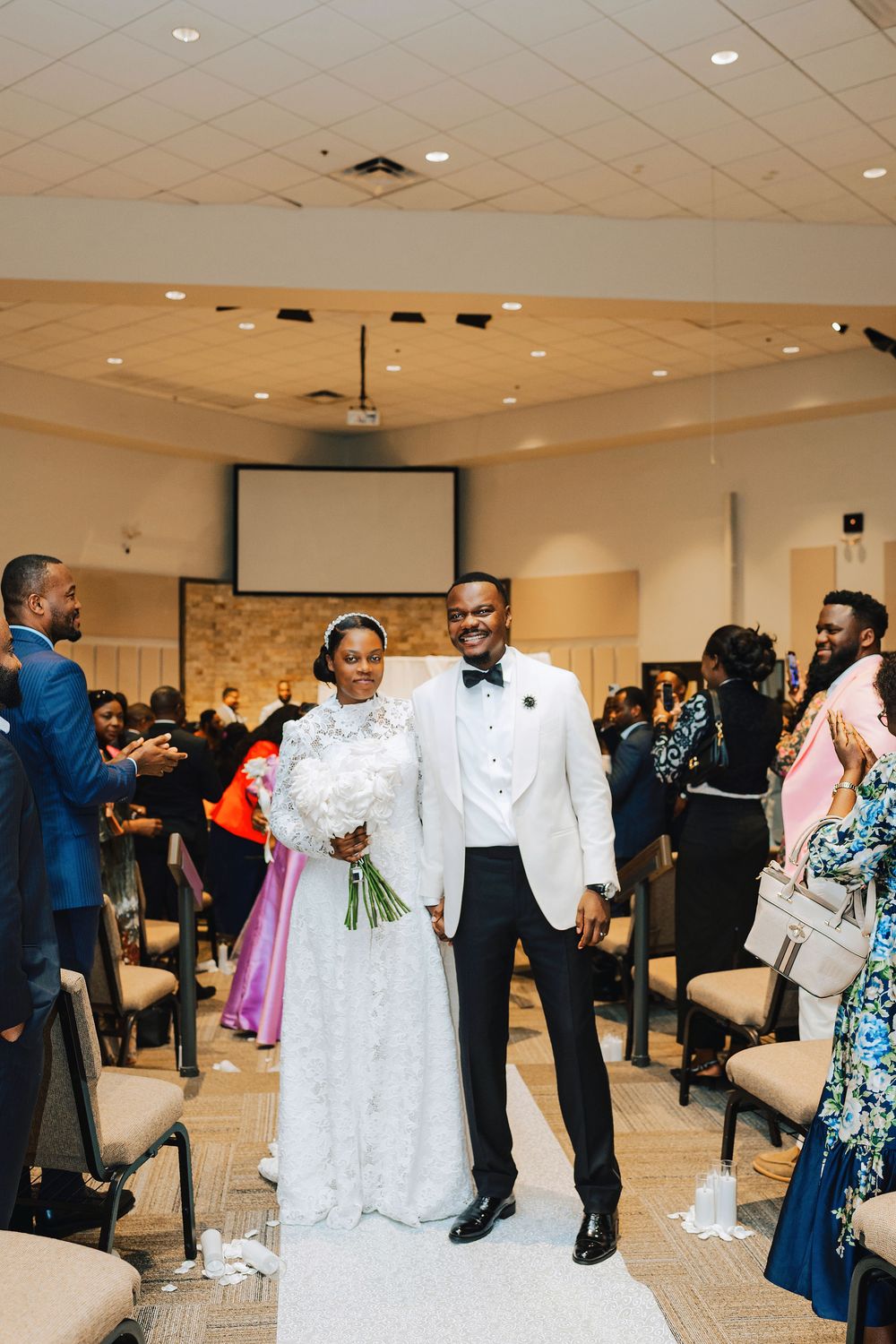 A joyful newlywed couple walks down the aisle in a church, bride in lace gown holding white bouquet, groom in white tuxedo.